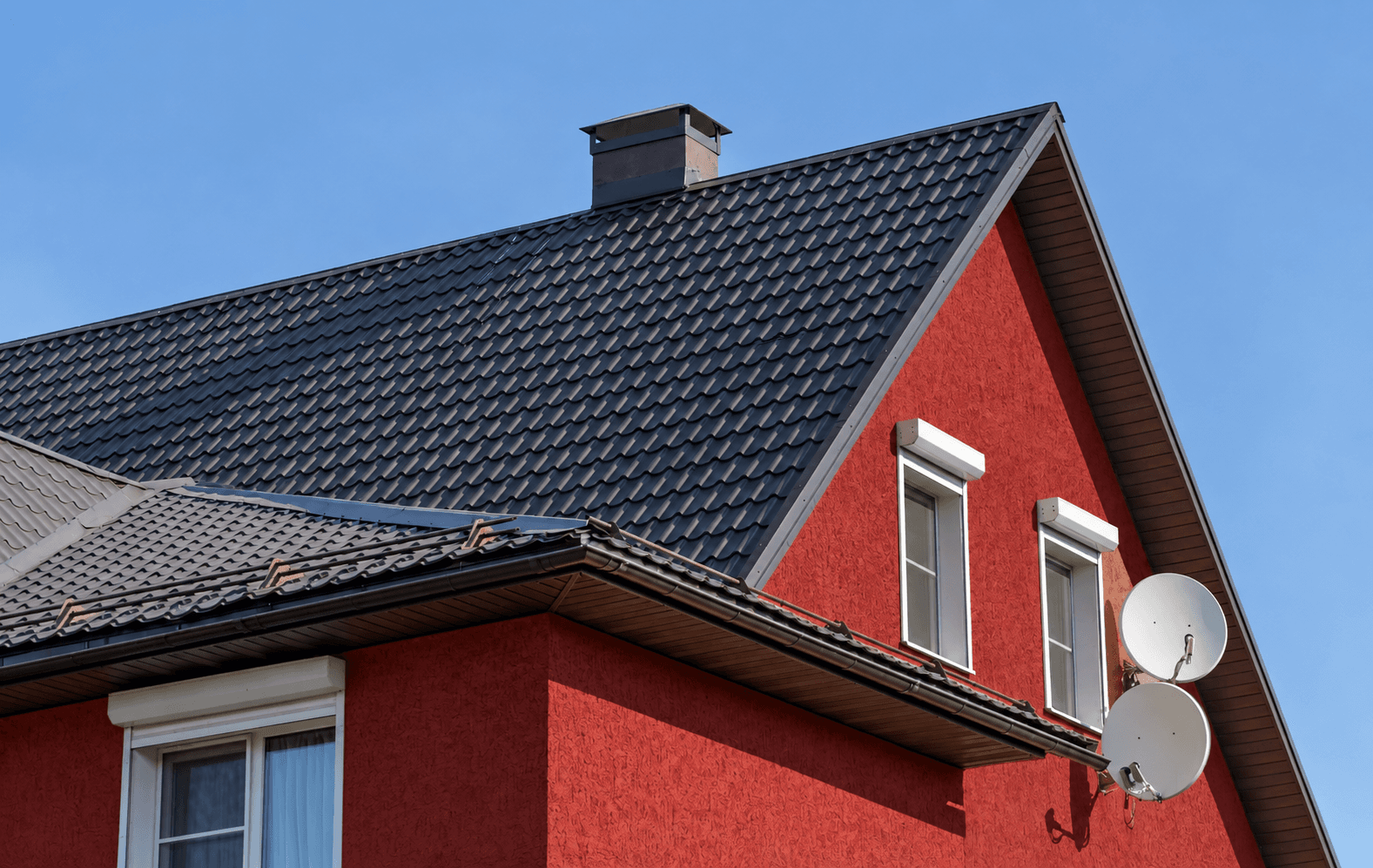 Steep dark metal tile gable above a deep red rendered facade, with a lower brown roof crossing beneath