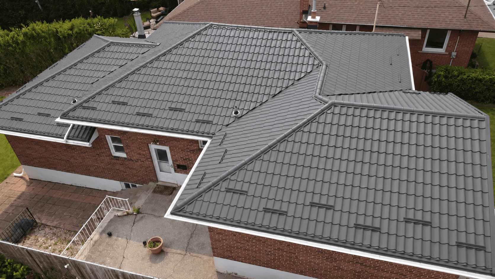 Grey metal tile roof stepped across low pitches above red brick walls, photographed from above