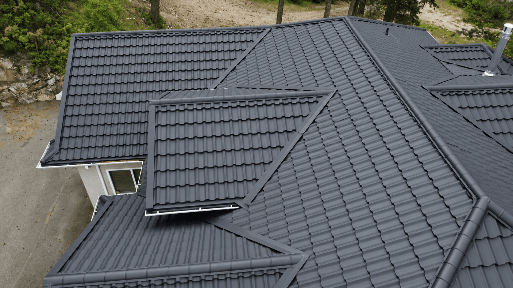 Complex charcoal metal tile roof of hips, valleys and a projecting gable on a rock-cut lot, seen from above